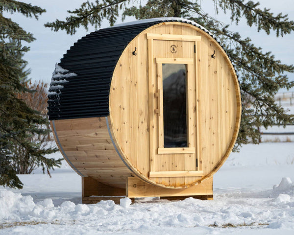 A cedar barrel sauna standing in snow in front of evergreens