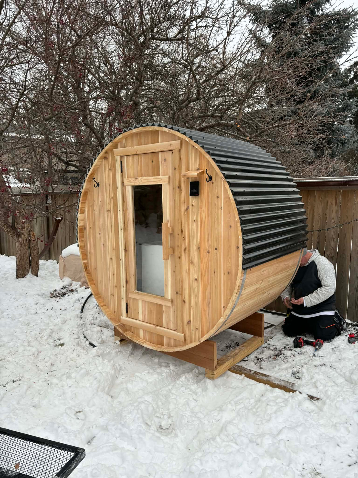 A cedar barrel sauna in a backyard with snow on the ground. RnR Hot Tubs and Spas