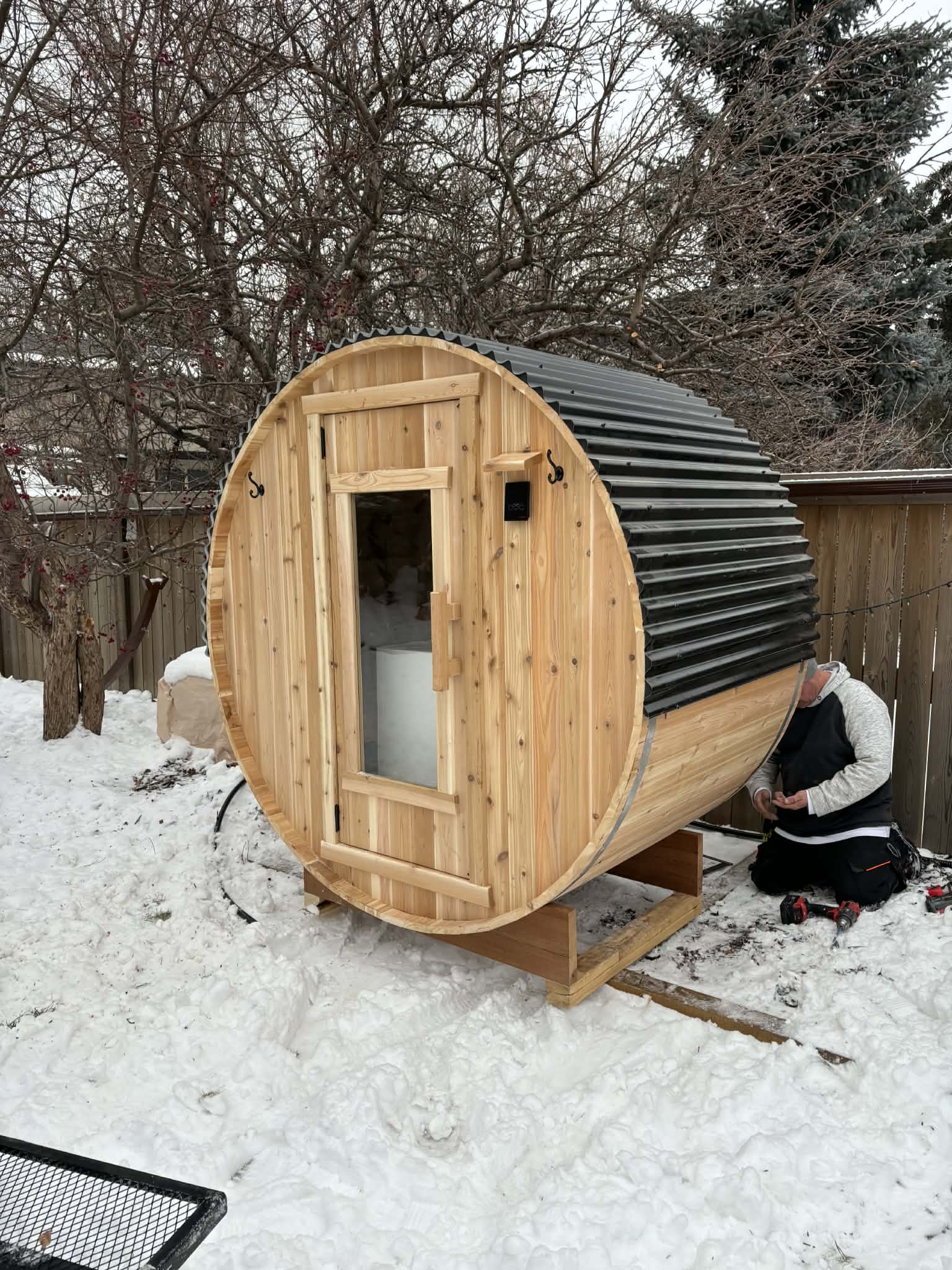 A cedar barrel sauna in a backyard with snow on the ground. RnR Hot Tubs and Spas